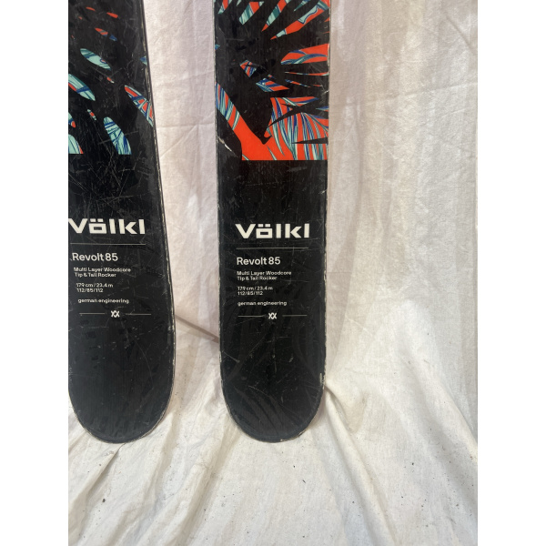 Close-up van de topsheet van twee Völkl Revolt 85 ski's (179 cm) met zichtbare specificaties: Multi Layer Woodcore, Tip & Tail Rocker, 112/85/112 mm sidecut, 23.4 m turnradius — tropisch blad-design in zwart, rood en turquoise