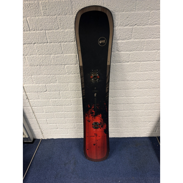 Komplettes Goodboards Reload Snowboard, schwarz mit braunen Rändern, rundem Logo, zwei Bindungsscheiben und rotem Ende, vor einer Backsteinwand