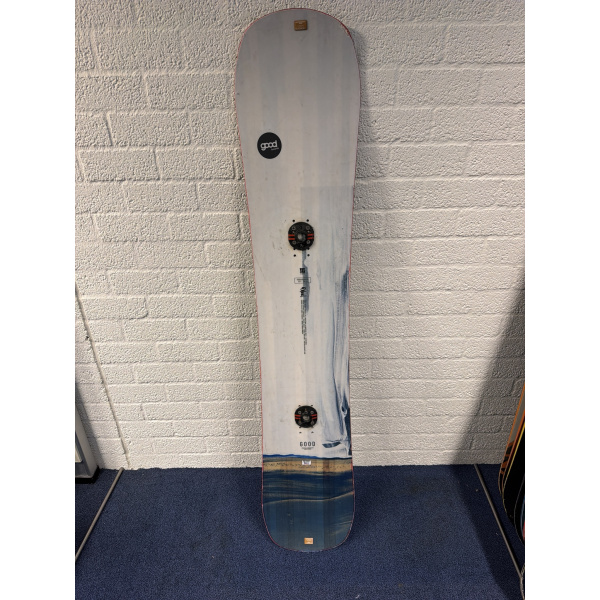 Goodboards Reload 67 XXWide Snowboard komplett von oben, weiß mit blauem Design und zwei Bindungsscheiben, vor einer weißen Backsteinwand